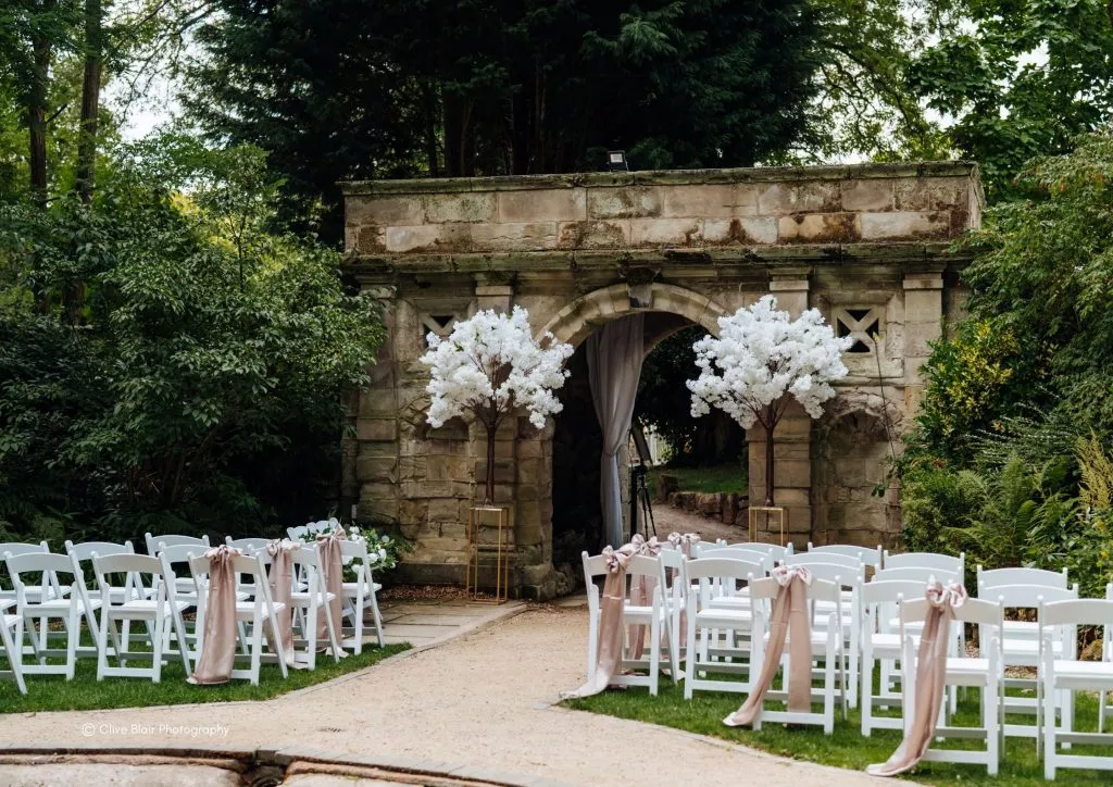 Outdoor wedding ceremony seating leading through a stone archway decorated with white blossom trees at Moor Hall Hotel & Spa.