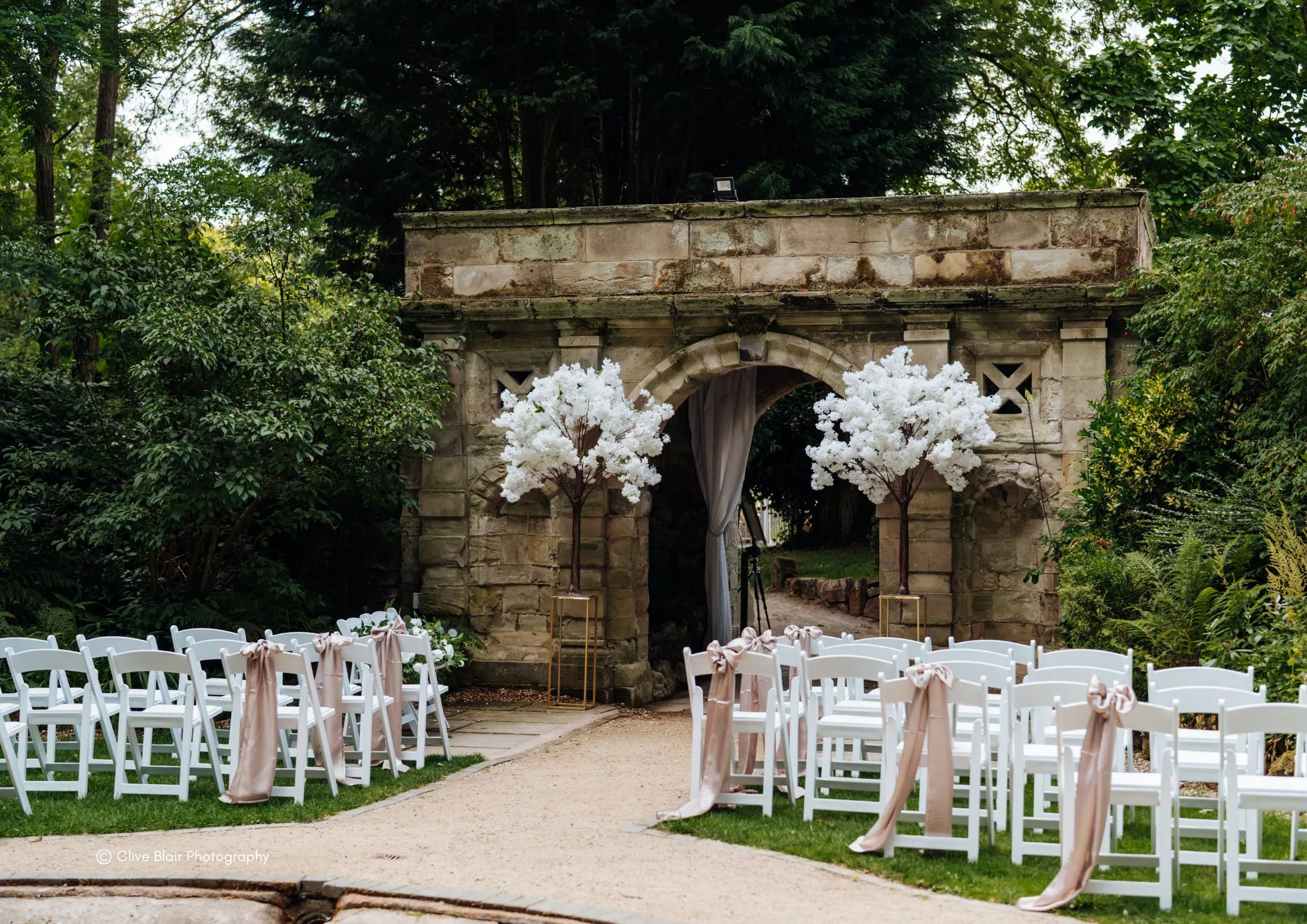 Outdoor wedding ceremony seating leading through a stone archway decorated with white blossom trees at Moor Hall Hotel & Spa.