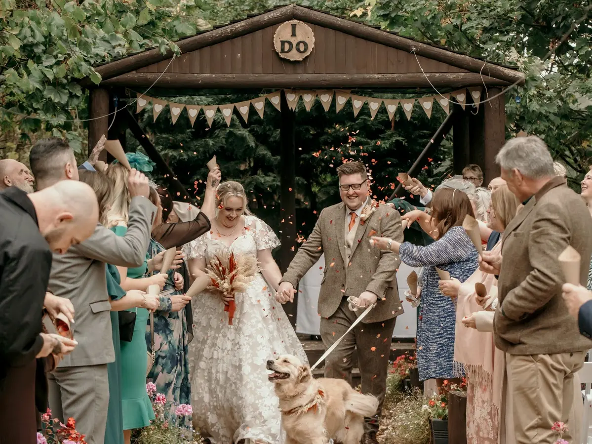 Bride and groom walking out of their wedding ceremony through a confetti shower with their golden retriever.